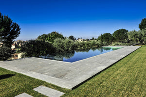 Piscine miroir en inox à débordement au ras de la plage en pierre, reflétant les arbres d'un jardin du Vaucluse